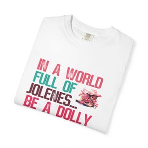 May include: A white t-shirt featuring the text "IN A WORLD FULL OF JOLENES... BE A DOLLY" printed in pink and teal. Next to the text is a graphic of a brown cowboy boot with a pink hat and pink flowers emerging from the top of the boot.
