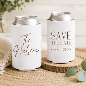 May include: Two white can coolers with brown text. One says "The Nielsens" and the other says "SAVE THE DATE 08.04.2027". The coolers are on a wooden coaster and a woven surface. Eucalyptus leaves are in the background.