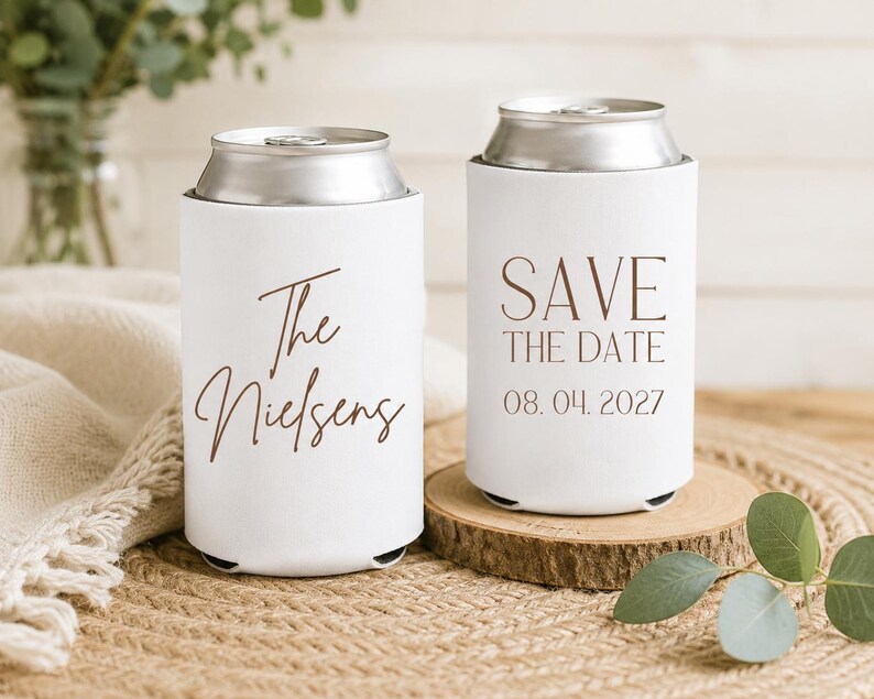 May include: Two white can coolers with brown text. One says "The Nielsens" and the other says "SAVE THE DATE 08.04.2027". The coolers are on a wooden coaster and a woven surface. Eucalyptus leaves are in the background.