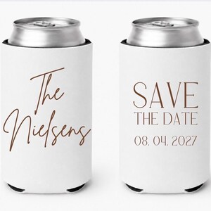 May include: Two white can coolers with brown text. One says "The Nielsens" in cursive, the other says "SAVE THE DATE 08.04.2027". The cans are silver and the coolers have black bottoms.