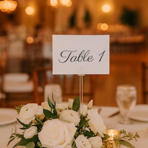 May include: A table centerpiece featuring a white floral arrangement with roses and greenery, a table number card that reads "Table 1", and lit candles in gold votive holders. The background is blurred, suggesting a wedding or event setting.