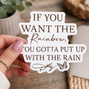 May include: A white die-cut sticker featuring the brown text IF YOU WANT THE Rainbow, YOU GOTTA PUT UP WITH THE RAIN. The design includes a signature script reading Dolly and a small butterfly illustration at the bottom right corner.