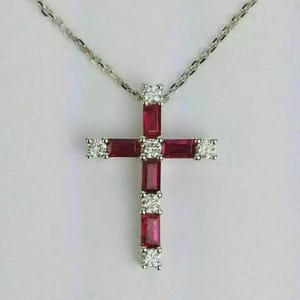 Peut inclure: Un collier argenté avec un pendentif en forme de croix. La croix est ornée de pierres rectangulaires rouge foncé taille baguette et de petites pierres rondes transparentes taille brillant, disposées en alternance sur les branches.