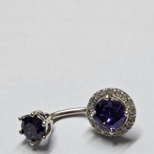 May include: A silver belly button ring with a purple gem surrounded by smaller clear gems. The other end of the ring has a smaller purple gem.