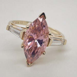 Op de afbeelding: Een gouden ring met een grote roze marquise-vormige edelsteen en twee kleinere baguette-vormige edelstenen aan de zijkanten.