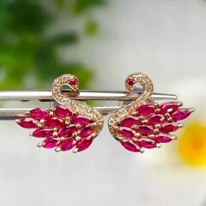 May include: A pair of gold swan earrings with red ruby stones. The swans are facing each other with their wings spread out.