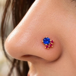 May include: A gold nose stud featuring a large, round, brilliant-cut blue gemstone accented by three smaller, round, vibrant pink gemstones arranged in a cluster formation, set securely in a polished gold metal prong setting.