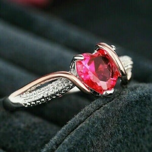 May include: A silver ring with a heart-shaped red gemstone and a twisted band. The band is accented with small clear stones.