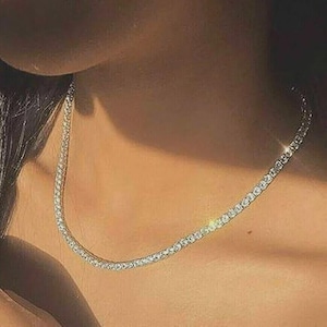 May include: A sparkling tennis necklace. The necklace features small, round, clear gemstones set closely together in a silver or white metal. The necklace is worn around the neck, reflecting light and creating a shimmering effect.