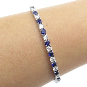 Puede incluir: Una pulsera de plata con piedras preciosas azules y blancas alternas. El diseño de la pulsera de tenis tiene piedras de corte redondo colocadas juntas. Las piedras preciosas están espaciadas uniformemente.