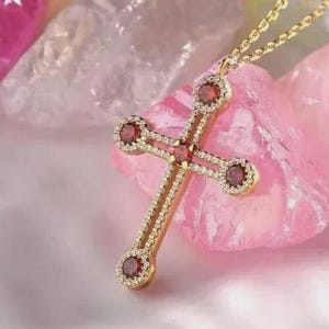 Könnte beinhalten: Eine goldfarbene Kreuz-Halskette mit vier runden, dunkelroten Steinen an den Enden und in der Mitte des Kreuzes. Der Rahmen des Kreuzes ist mit kleinen, klaren Pavé-Steinen besetzt und hängt an einer zierlichen goldfarbenen Kette.