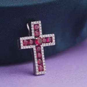 May include: A cross pendant featuring a central row of round, deep red rubies framed by a border of small, clear white stones. The metallic setting provides a structured outline for the gemstones, creating a distinct contrast between the red and white.