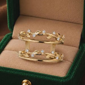 Puede incluir: Un anillo de doble banda en tono dorado con un delicado motivo de enredadera y hojas. Las dos bandas están conectadas por barras verticales y las hojas están acentuadas con pequeñas piedras claras y brillantes a lo largo de los bordes.