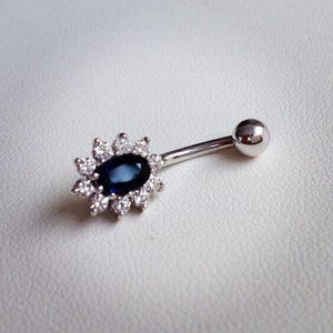 May include: A silver belly button ring with a blue sapphire gemstone surrounded by a halo of smaller clear gemstones.