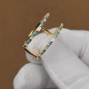 May include: A gold open-style ring featuring two parallel bands connected by a central bar. Each band is set with alternating clear baguette-cut stones and small round turquoise-colored accents, creating a symmetrical and geometric design.