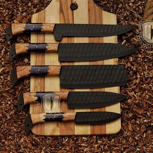 May include: A set of five black chef knives with wooden handles and dark blue accents, displayed on a wooden cutting board. The blades have textured surfaces. The handles are a light brown color.