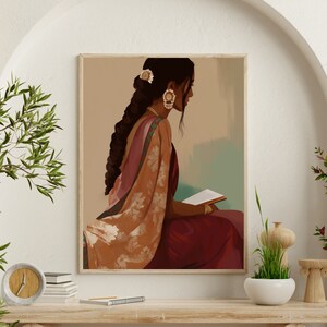 May include: A framed art print showing a woman in traditional Indian dress, reading a book. The artwork uses burgundy, peach, and gold tones, with detailed clothing and jewellery. The woman has long, dark hair and wears earrings and a hair accessory. The print is displayed on a wooden table.