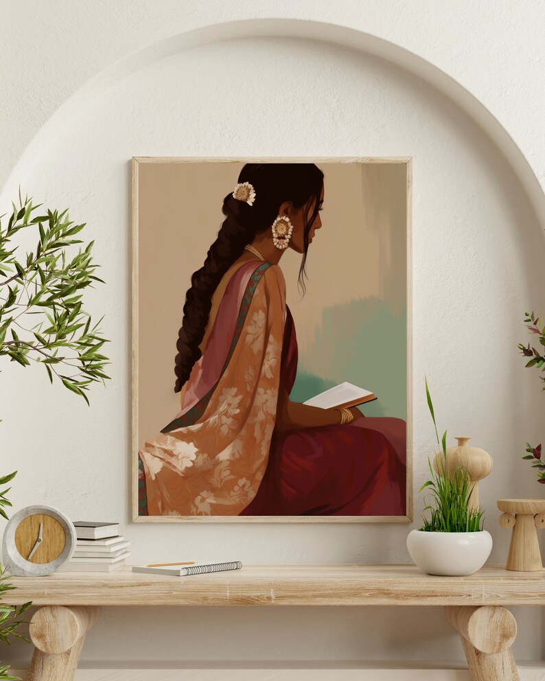 May include: A framed art print showing a woman in traditional Indian dress, reading a book. The artwork uses burgundy, peach, and gold tones, with detailed clothing and jewellery. The woman has long, dark hair and wears earrings and a hair accessory. The print is displayed on a wooden table.