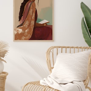May include: A framed art print showing a person in Indian dress, reading. The artwork uses burgundy, peach, and gold tones. The print is in a light wooden frame, against a neutral wall. A woven chair and cushion are in the foreground, with a blanket draped over the chair.