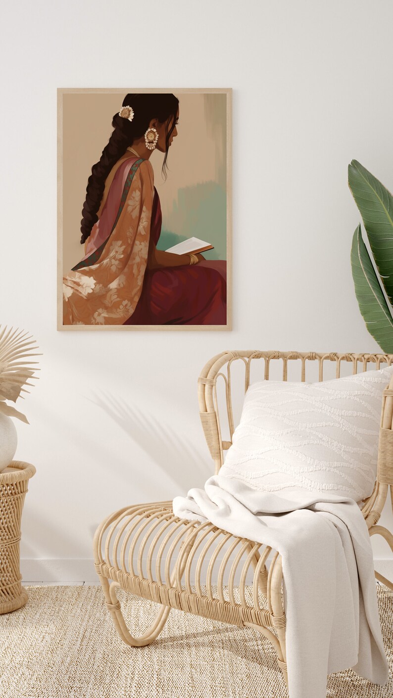 May include: A framed art print showing a person in Indian dress, reading. The artwork uses burgundy, peach, and gold tones. The print is in a light wooden frame, against a neutral wall. A woven chair and cushion are in the foreground, with a blanket draped over the chair.