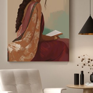 May include: An art print showing a woman in traditional Indian dress, seated and reading a book. The artwork uses burgundy, peach, and gold tones, with detailed jewellery and clothing. The print hangs above a white armchair.