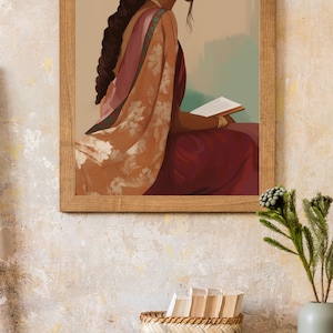 May include: A framed print shows a woman in a sari and jewellery, reading a book. The artwork is in a wooden frame. A woven basket, a small round box, and a gold clock sit on a wooden table, with a vase of greenery.