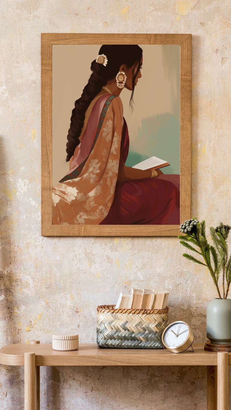 May include: A framed print shows a woman in a sari and jewellery, reading a book. The artwork is in a wooden frame. A woven basket, a small round box, and a gold clock sit on a wooden table, with a vase of greenery.