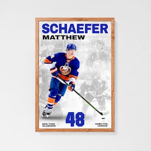 May include: A framed sports poster featuring hockey player Matthew Schaefer in a blue and orange New York Islanders jersey, holding a hockey stick. The text reads SCHAEFER MATTHEW, 48, NEW YORK ISLANDERS, and HAMILTON, CANADA, with a faded background.