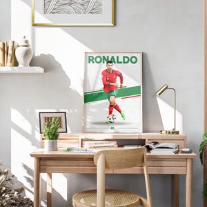 May include: A framed poster featuring Cristiano Ronaldo in a red soccer uniform with green and white accents. The word "RONALDO" is printed in green at the top. The poster is displayed on a wooden desk with a lamp and a small plant.