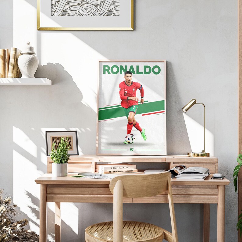 May include: A framed poster featuring Cristiano Ronaldo in a red soccer uniform with green and white accents. The word "RONALDO" is printed in green at the top. The poster is displayed on a wooden desk with a lamp and a small plant.