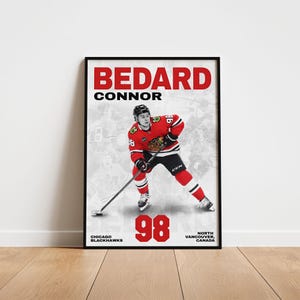 Może przedstawiać: Oprawiony plakat z hokeistą Connorem Bedardem w stroju Chicago Blackhawks. Plakat ma czarną ramę i zawiera nazwisko zawodnika, numer 98 oraz informacje o drużynie. Tło przedstawia wyblakły obraz innych graczy.