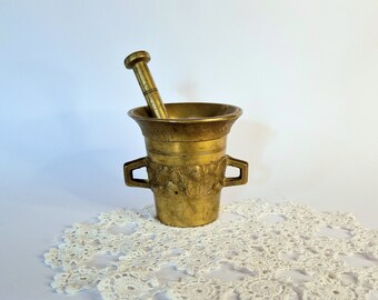 May include: A brass mortar and pestle set with an ornate design. The mortar has two handles and the pestle is smooth. The set is sitting on a white crocheted doily.