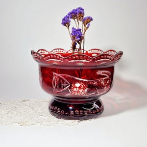 Könnte beinhalten: Eine rubinrote Glasschale mit gewelltem Rand und Sockel. Die Schale hat geätzte Muster und ist mit getrockneten lila Blumen gefüllt. Ein dekoratives Objekt.