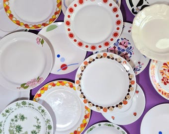 Może przedstawiać: Kolekcja białych ceramicznych talerzy z różnymi kolorowymi wzorami. Niektóre talerze mają wzory kwiatowe, inne geometryczne lub abstrakcyjne wzory w odcieniach czerwieni, pomarańczy, żółci i zieleni. Talerze ułożone są na fioletowej powierzchni.