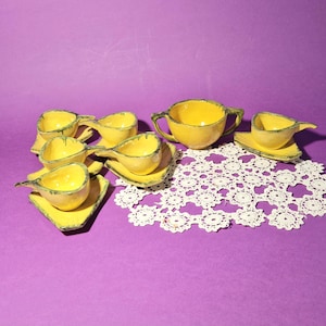 Może przedstawiać: Zestaw żółtych ceramicznych filiżanek i spodków z zielonymi obwódkami. Zestaw zawiera cukiernicę i dzbanuszek do śmietanki. Filiżanki mają unikalny, wydłużony kształt. Zestaw jest ułożony na fioletowej powierzchni z białą koronkową serwetką.