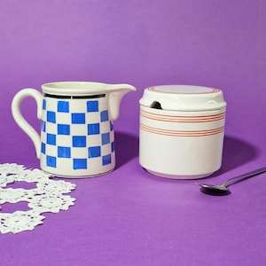 May include: A white ceramic creamer with a blue chequered pattern and a matching sugar bowl with red stripes. A small silver spoon rests beside the sugar bowl. The items are on a purple surface.