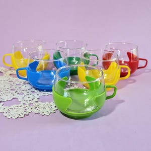 May include: Six clear glass teacups with colorful silicone sleeves and handles. The sleeves and handles are in yellow, blue, green, and red. The cups are arranged on a purple surface with a white lace doily.