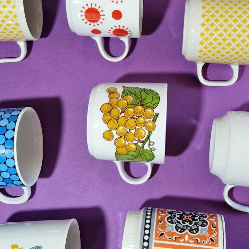Może przedstawiać: Zdjęcie z g&oacute;ry kilku białych ceramicznych kubk&oacute;w z r&oacute;żnymi kolorowymi wzorami, w tym winogronami, geometrycznymi wzorami i motywami kwiatowymi. Kubki są ułożone na jednolitym fioletowym tle, prezentując ich unikalne style.