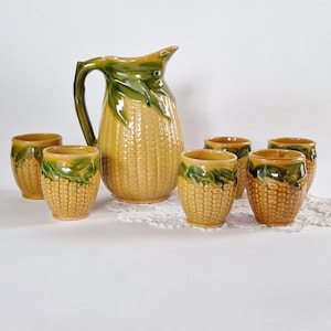 Op de afbeelding: Een set van zes gele en groene keramische bekers en een kan in de vorm van maïs. De bekers en de kan hebben een getextureerd oppervlak dat lijkt op maïskorrels.