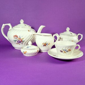 Könnte beinhalten: Ein cremefarbenes Teeservice mit floralen Designs. Das Set umfasst eine Teekanne, eine Zuckerdose, einen Sahnekännchen, eine Teetasse, eine Untertasse und eine Servierschale mit Deckel. Die Teile haben eine Spiralstruktur und goldfarbene Verzierungen.