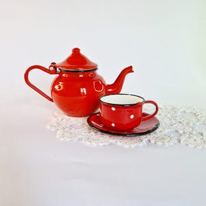 Puede incluir: Tetera y taza de té de esmalte rojo con lunares blancos. La tetera tiene una tapa y un pico. La taza tiene un platillo.