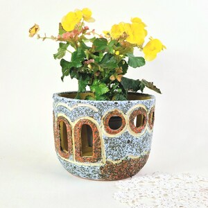 May include: A ceramic planter with a textured light blue and brown speckled finish featuring arched and circular cutouts along the sides. The planter contains a green plant with bright yellow flowers blooming from the top opening.
