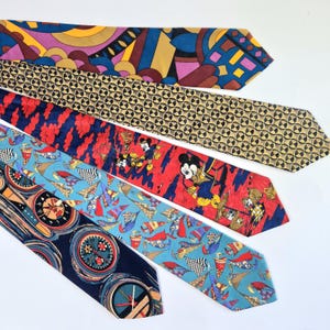 Op de afbeelding: Vijf kleurrijke stropdassen met verschillende patronen worden getoond. De stropdassen hebben ontwerpen zoals geometrische vormen, cirkels en een Mickey Mouse-print. Kleuren zijn onder meer blauw, geel, rood en bruin.