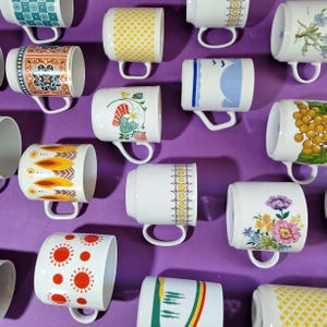 Może przedstawiać: Kolekcja ceramicznych kubk&oacute;w z r&oacute;żnymi wzorami i wzorami. Kubki mają motywy kwiatowe, geometryczne i abstrakcyjne w kolorach takich jak ż&oacute;łty, pomarańczowy, niebieski i czerwony. Każdy kubek ma biały uchwyt i jest umieszczony na fioletowym tle.