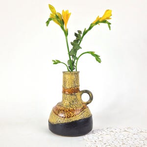 May include: A ceramic vase with a bulbous base and a narrow neck featuring a side handle. The surface has a textured yellow and brown mottled glaze with a solid dark brown band at the bottom. Three yellow freesia flowers are inside the vase.