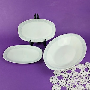 Op de afbeelding: Drie witte keramische schalen met een lichtgrijs gestructureerd randpatroon. De set bevat één ovale schaal op een zwarte standaard, één kleiner ovaal bord en één grote ovale kom, allemaal met een consistent randontwerp.