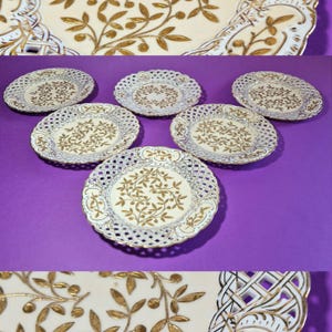 Può includere: Un set di sei piatti in porcellana bianca e oro con un delicato design traforato. I piatti presentano un motivo botanico dorato al centro e sui bordi. I piatti sono disposti su una superficie viola, con un vassoio da portata abbinato visibile.