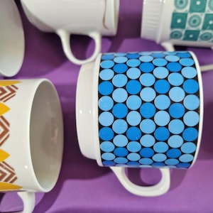 Może przedstawiać: Kolekcja białych ceramicznych kubk&oacute;w z r&oacute;żnymi wzorami. Jeden kubek ma niebieski wz&oacute;r w kropki, a inne geometryczne i kwiatowe wzory. Kubki mają białe uchwyty i są ułożone na fioletowej powierzchni.