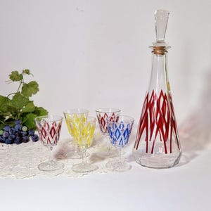 Puede incluir: Una jarra de vidrio vintage con tapón transparente y diseño de rombos rojos, junto con cuatro copas de vino a juego en rojo, amarillo y azul. Cristalería para servir vino.