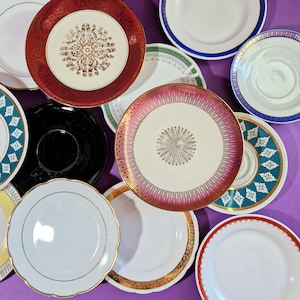 Op de afbeelding: Een verzameling vintage borden en schotels in verschillende kleuren en ontwerpen. Inclusief witte, zwarte, rode, gouden en turquoise borden met bloemen- en geometrische patronen. Sommige borden hebben gouden randen, andere blauwe of groene accenten.